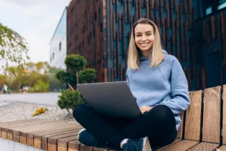 Duales Studium in der Nähe - junge Frau auf einer Bank in der Stadt, lächelt in die Kamera und hat den Laptop auf dem Schoß.