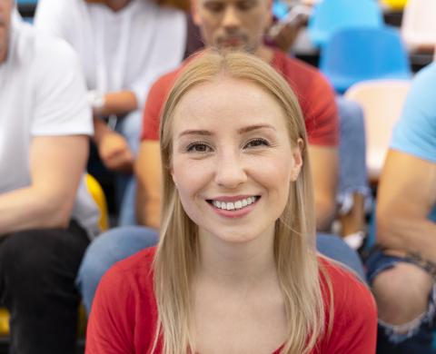 Blonde Frau in rotem Shirt, lächelt. Im Hintergrund sind weitere Personen zu sehen.